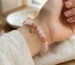 Minimalist rose quartz self compassion bracelet worn on a wrist, featuring faceted pink crystal beads, gold spacers, and a delicate heart-shaped dangling charm with a crystal center.
